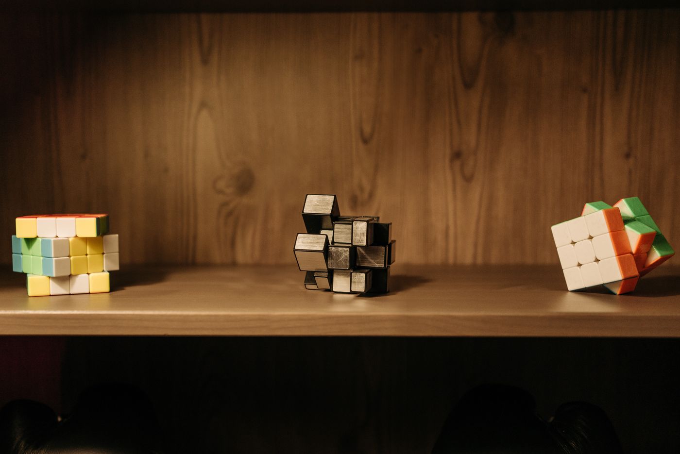 Three different puzzle cubes standing side by side on a wooden shelf